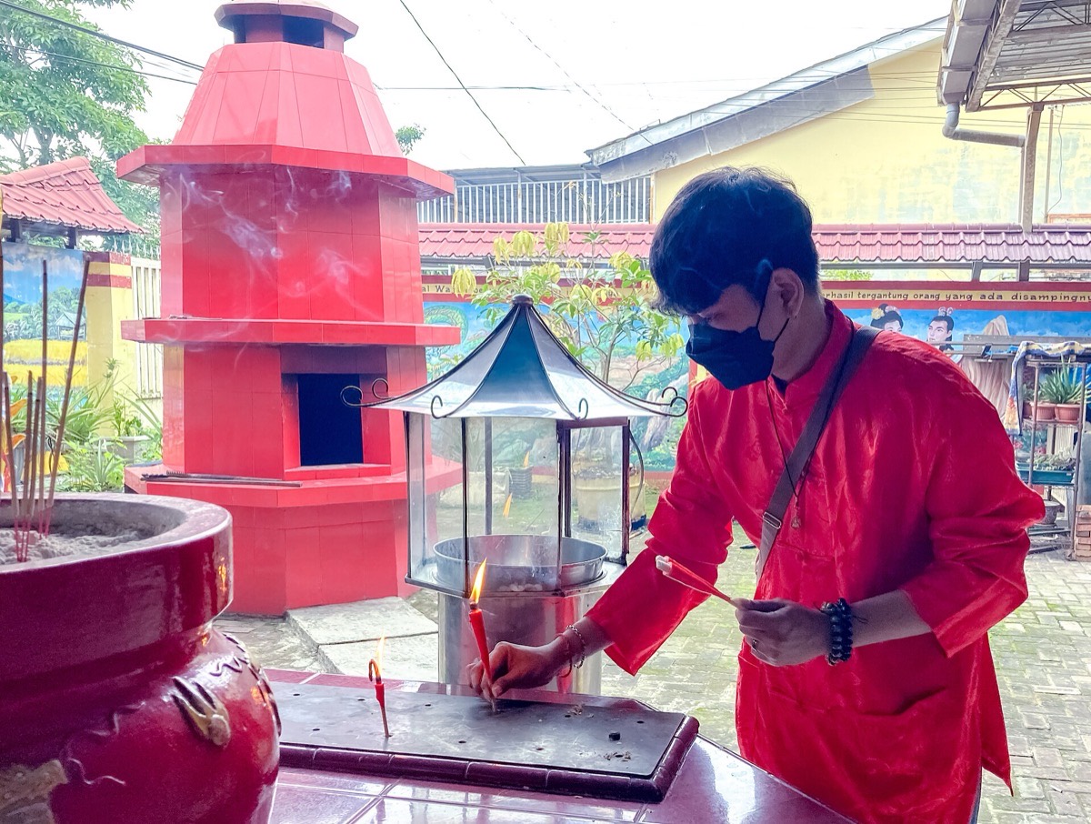 A man in a red shirt bending to light a thin red candle on a temple candle stand, beside a steel wind shelter and a censer full of incense sticks.
